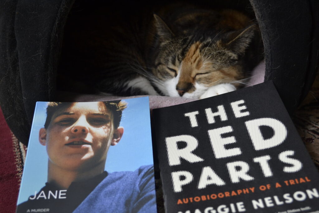 A calico tabby sleep peacefully in a domed bed beside two books by Maggie Nelson.