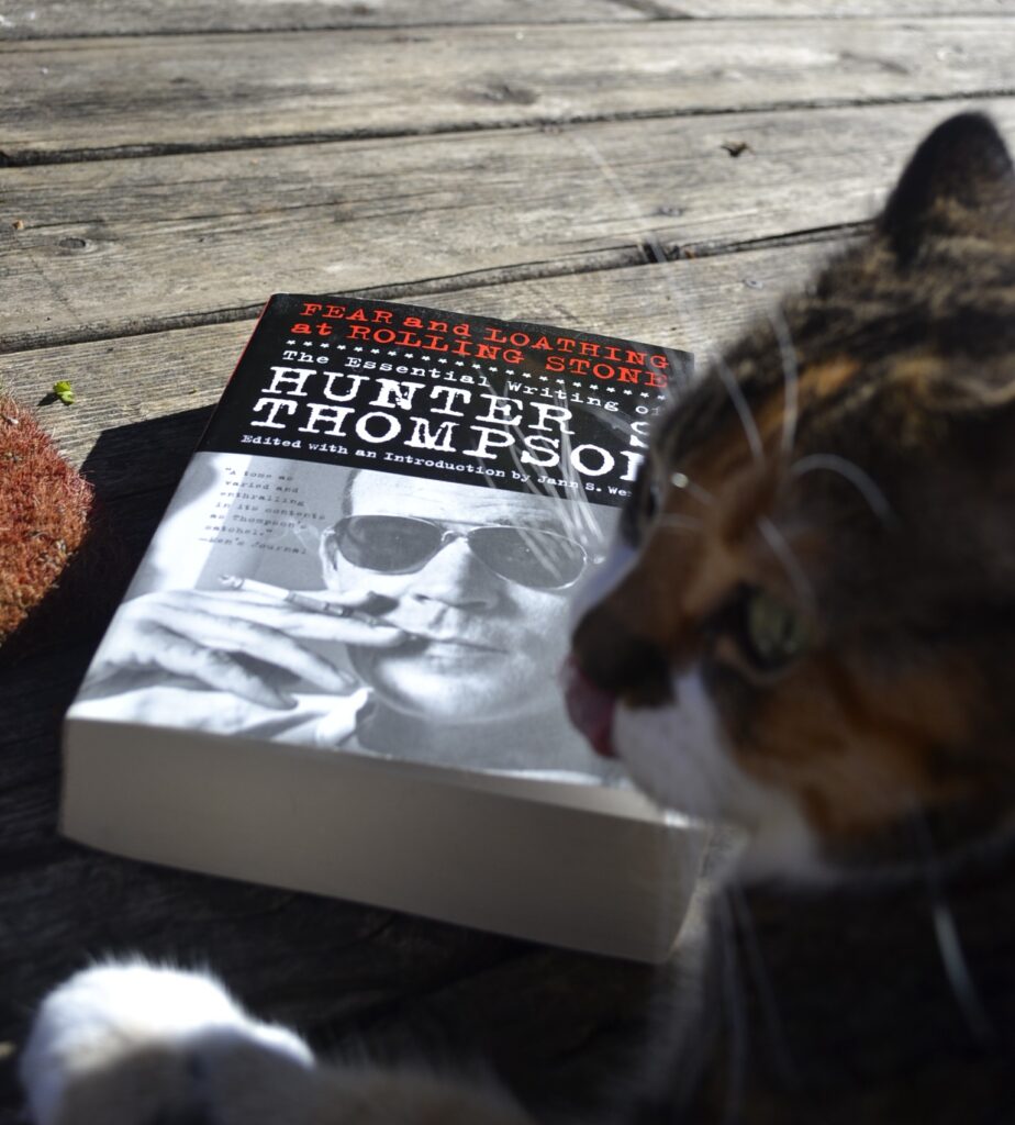 A calico tabby licks her nose in front of a thick paperback book.